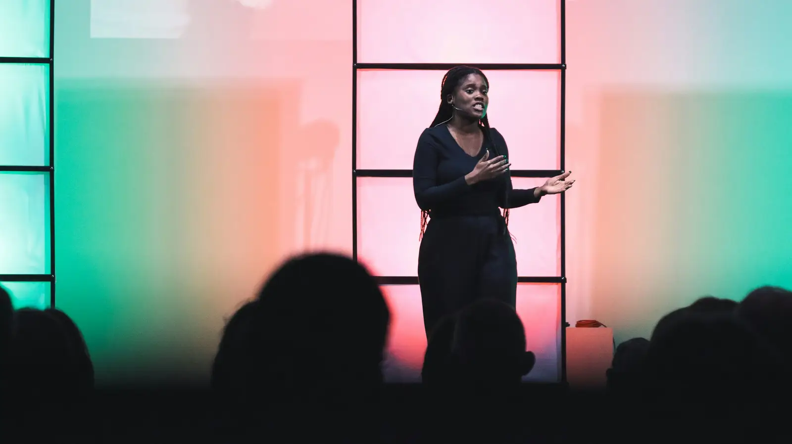 A woman speaks on a stage, gesturing as she explains her idea, while the audience listens in silhouette.