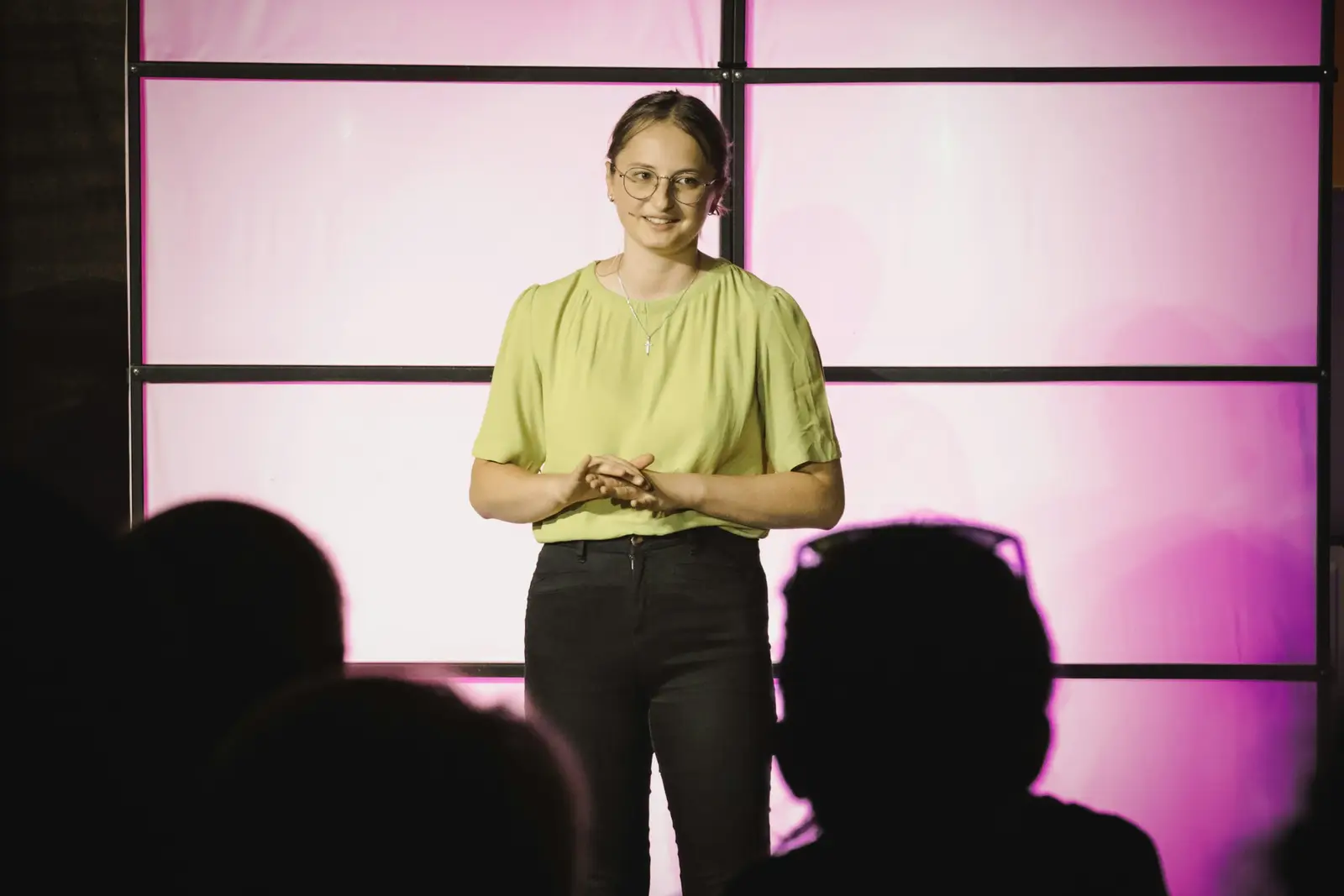 A woman in a green top presents on a stage lit in pink, with the audience in the foreground.